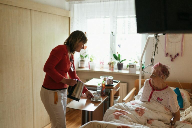 Older woman in hospital bed with younger woman standing next to bed assisting with pouring a drink for her.
